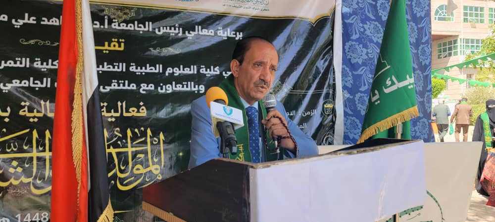 Man in a blue suit speaks at a podium during a formal event, with an Arabic banner and flags behind him.