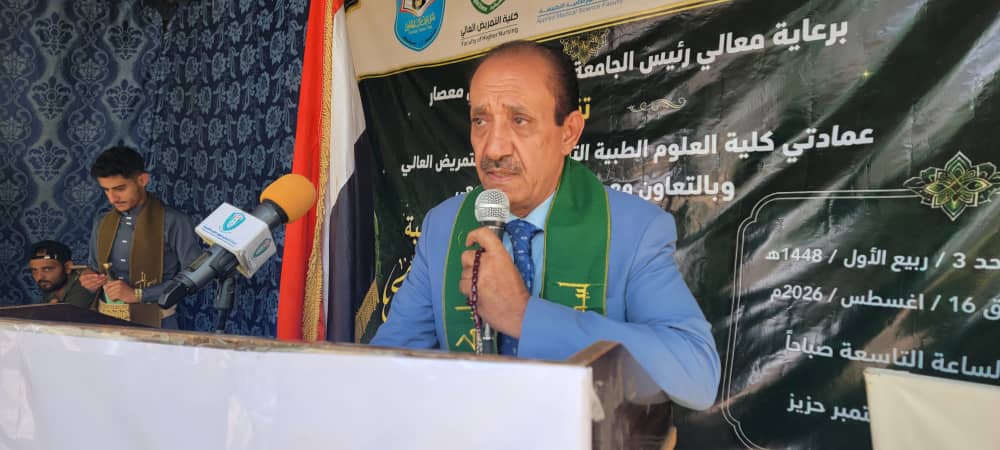 Man in a blue suit speaks into a microphone at a podium during an event, with a green scarf and Arabic banner behind him.