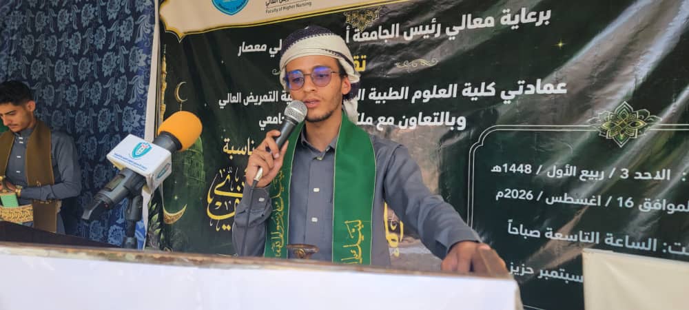 Young man wearing a white headband and green scarf speaks into a microphone at a podium during a formal event, with an Arabic banner behind him.