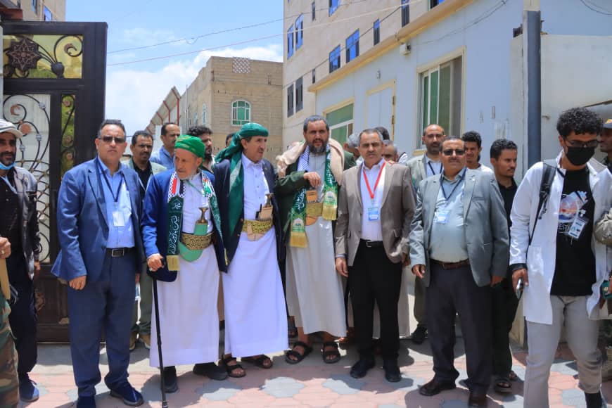 Group of men in traditional white robes and green sashes posing in a street, some wearing lanyards, during a ceremony or gathering.