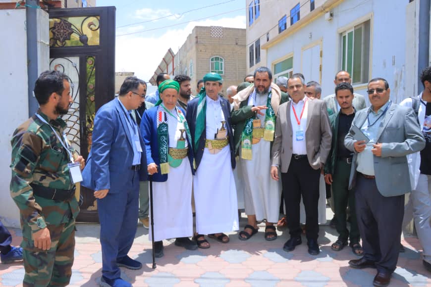 Group of men, some in traditional white robes and green turbans, posing together outside a building on a sunny day.