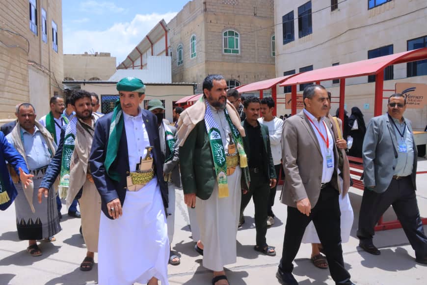 Group of men in traditional Yemeni clothing and green scarves walking together in a sunlit courtyard with brick buildings in the background.