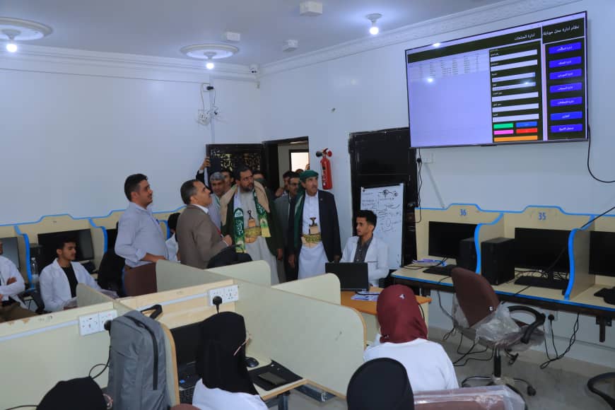 Group of people in traditional attire gathered around a desk in a computer lab as a presenter explains something at a laptop.