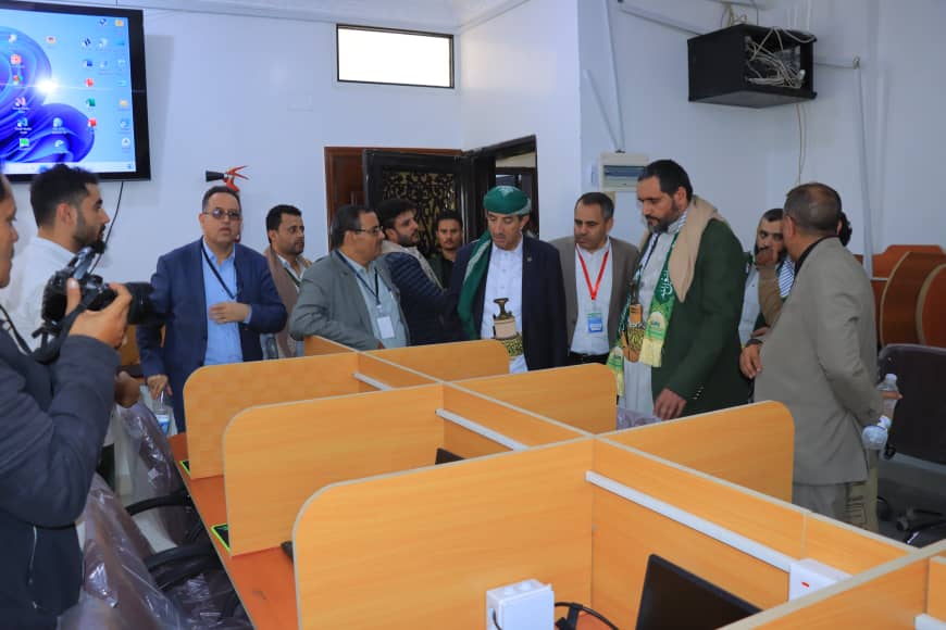 Group of men in business attire touring a room with wooden cubicles; a photographer takes their picture in the foreground.