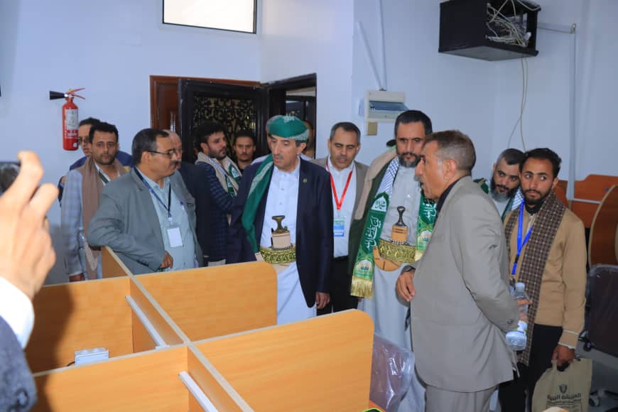 Group of men in suits and traditional dress touring a room with wooden cubicle desks.