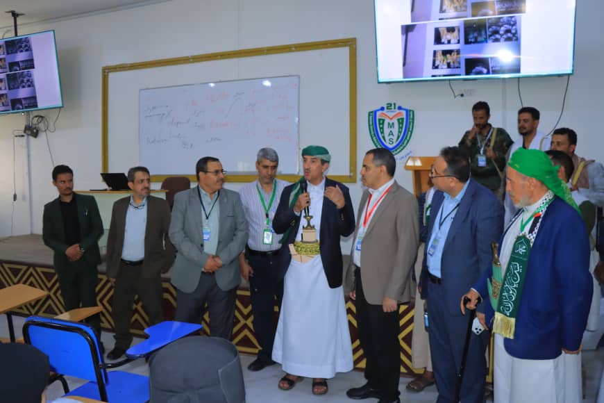 Group of men in suits and traditional dress on a stage, a cleric holding a microphone at center addressing the audience, with a whiteboard and screens in the background.