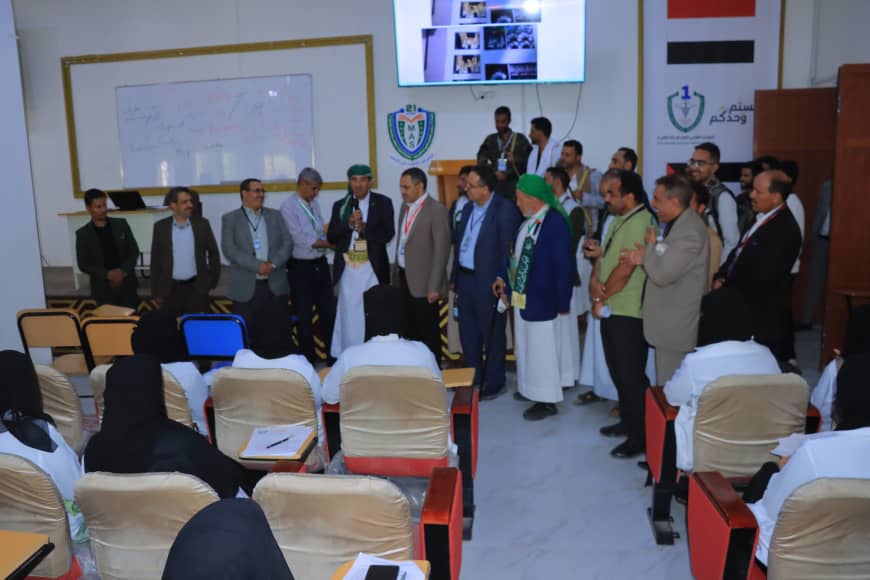 Group of men in suits and traditional dress standing at the front of a conference room, addressing an audience seated in rows.
