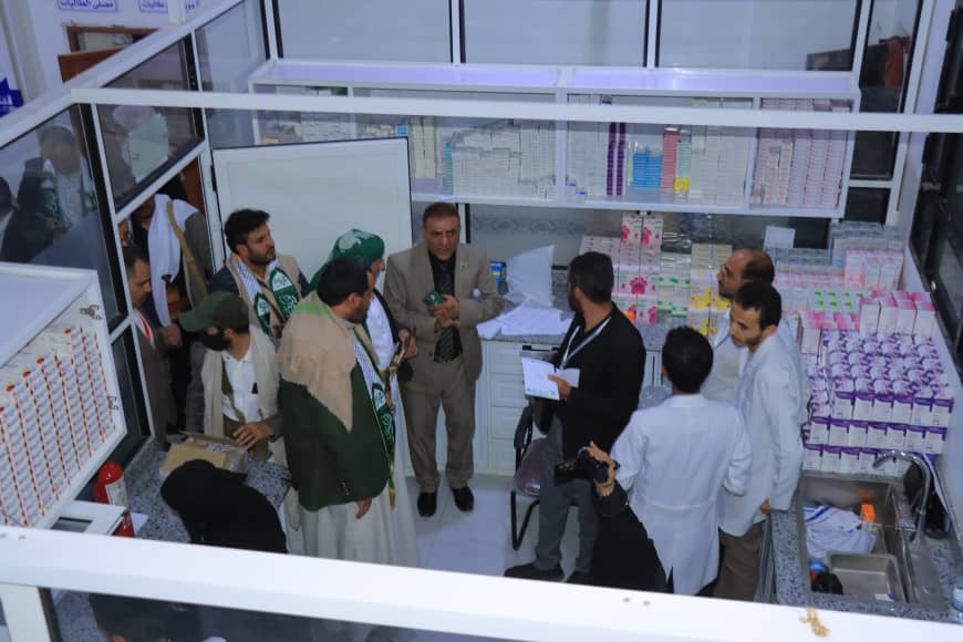 Group of men, some in traditional attire, stand in a pharmacy discussing a document held by a man in a dark shirt; shelves stocked with medicines surround them.