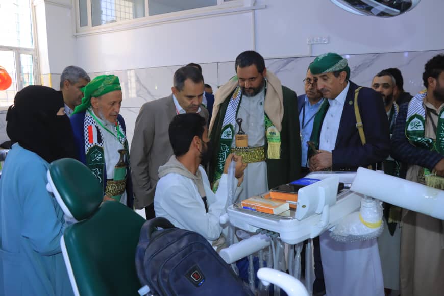 Dentist chair in a clinic with a patient being examined while a group of men in green scarves observe nearby.