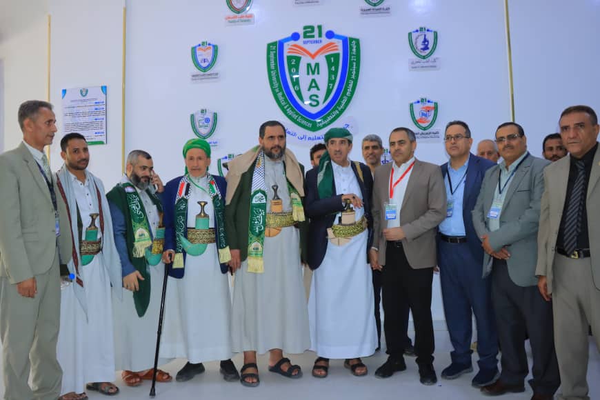 Group of men in traditional white robes and suits pose in front of a MAS event backdrop with a large green MAS emblem and badges nearby.