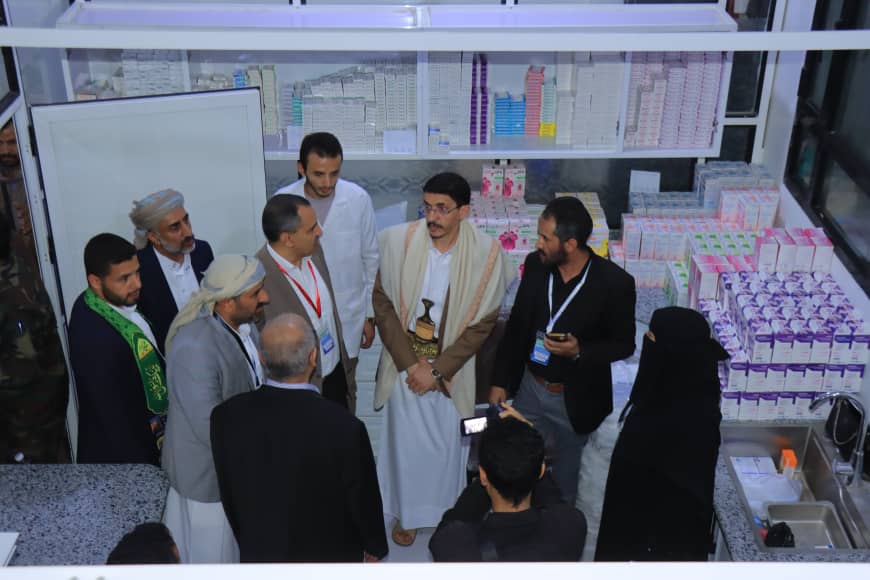 Group of men, some in traditional robes, gather in a pharmacy showroom, standing around and likely discussing amid shelves of medicine boxes behind them.