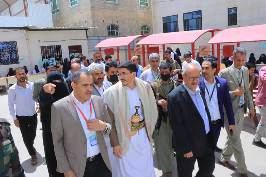Group of men in suits and traditional clothing walking together at an outdoor event, several wearing lanyards, with red canopies and a building in the background.