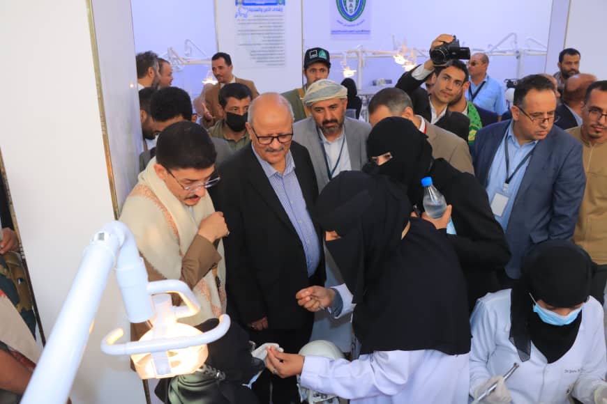 Crowd of people at a medical demonstration; a woman in a black hijab hands a small item to a suited man while others observe.