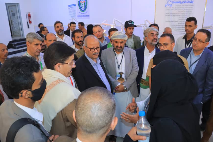 A woman in a black abaya speaks to a group of men in suits and traditional robes at an indoor event, attendees wearing lanyards.
