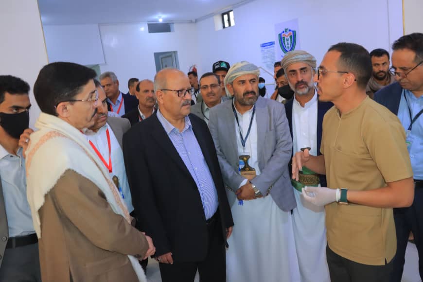 Group of men in a hallway listening to a young man in a tan shirt presenting a small crafted item to the crowd, some wearing traditional clothing and lanyards.