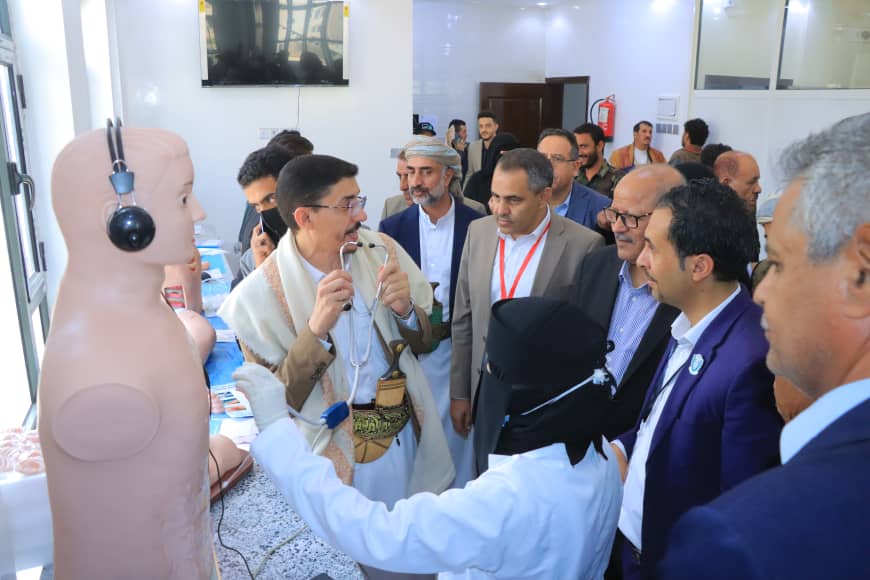 Medical demonstration: a group of professionals observe a doctor using a stethoscope beside a life-size mannequin wearing a headset in a clinic-like setting.