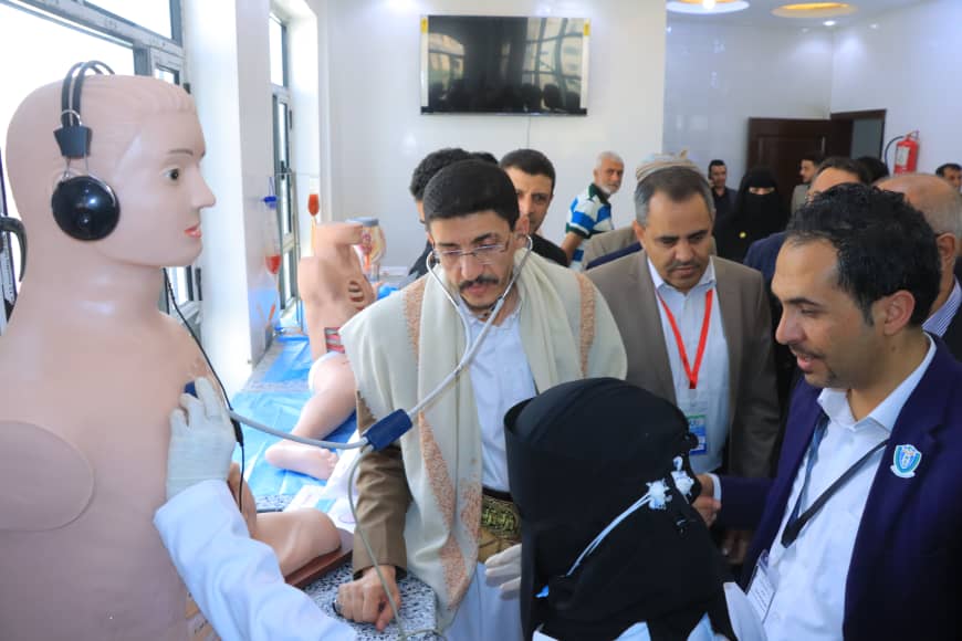 Group of professionals observes a medical training on a life-size patient mannequin connected to monitors and headphones in a bright room.