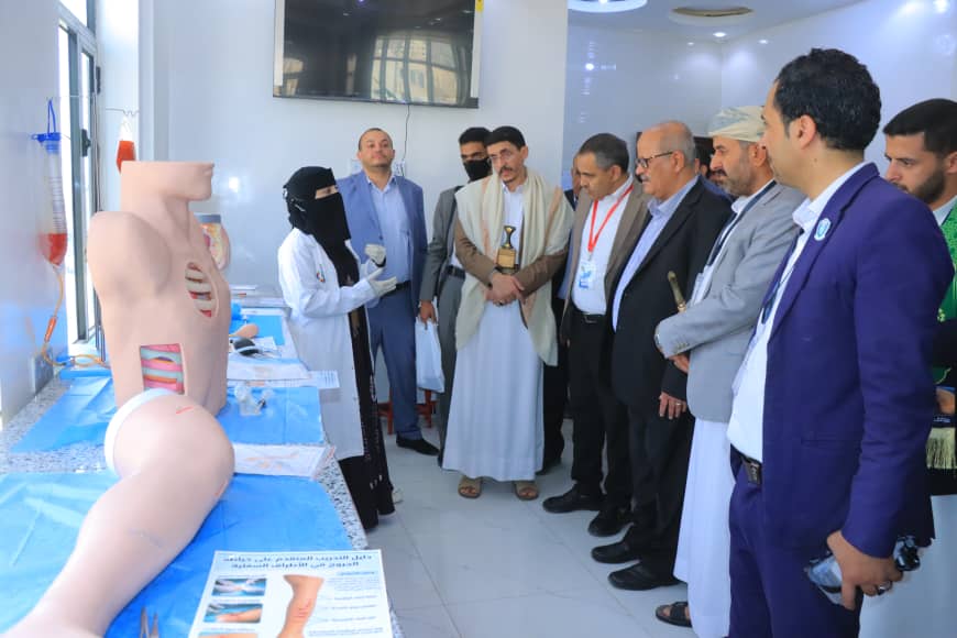 Group of professionals inspect a medical training exhibit with a large anatomical torso and leg models on blue tables.