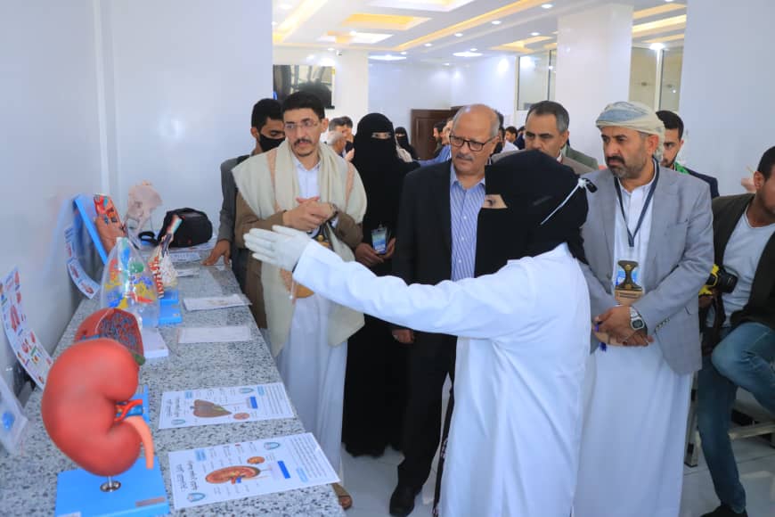 Physician in white coat and gloves explains organ models to visitors at a medical expo, with a kidney model on the counter nearby.