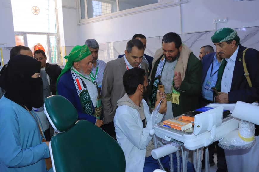 Group of men in traditional and business attire around a clinician at a dental chair, discussing equipment in a clinic setting.