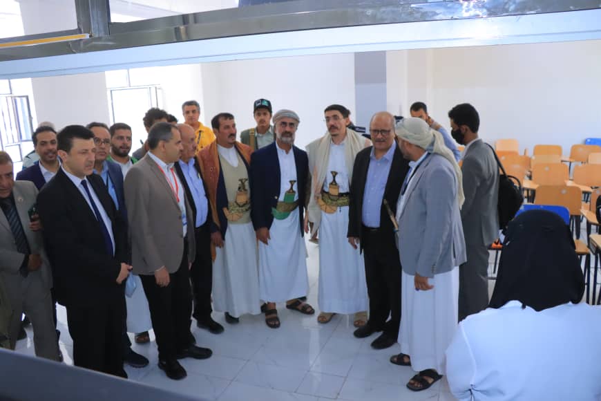Group of men in traditional robes and suits standing in a classroom or lecture hall, forming a circle for a gathering or presentation.