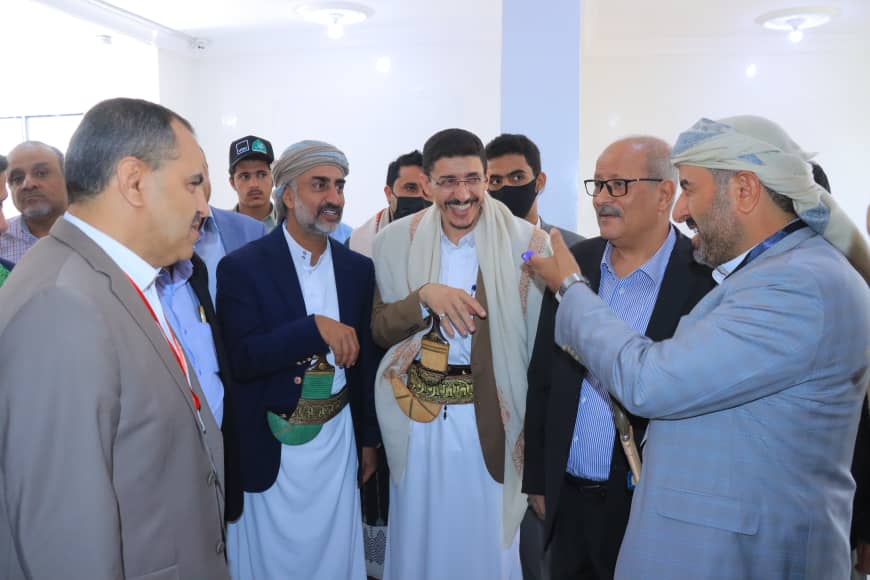 Group of men in traditional outfits and suits talking indoors at an event, one man gesturing with his hands as others listen.