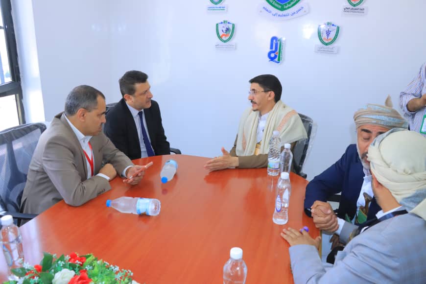 Five men sit around a wooden conference table in a discussion, water bottles on the table and badges visible on their lanyards.