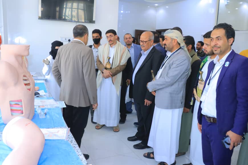 Group of men in traditional robes and suits stand in a bright room, observing an educational medical display of life-size torso models with exposed organs on a blue table(s).