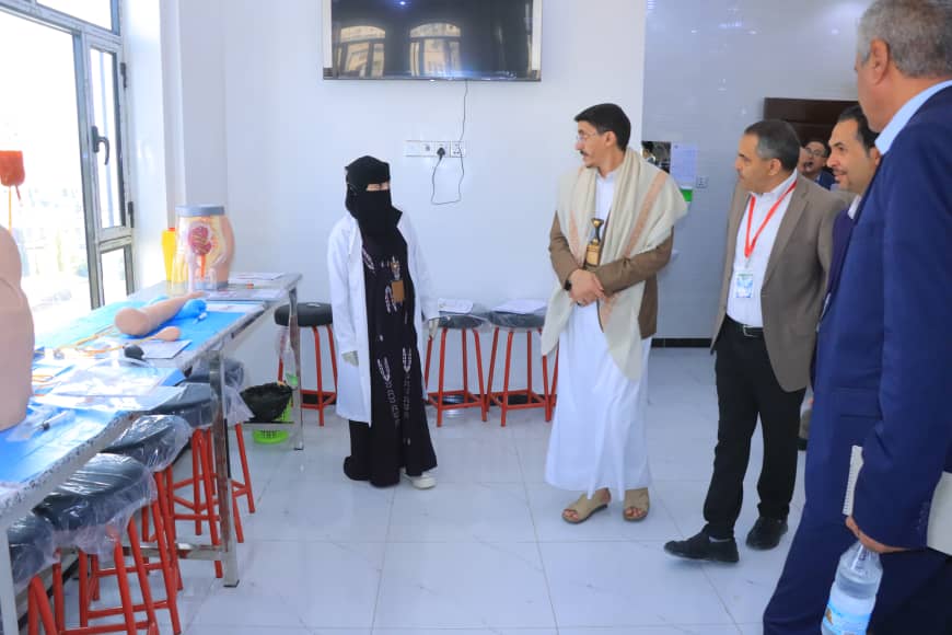 Visitors watch a medical training demonstration in a bright clinic room, with a woman in a niqab and a man in traditional robes at the center.