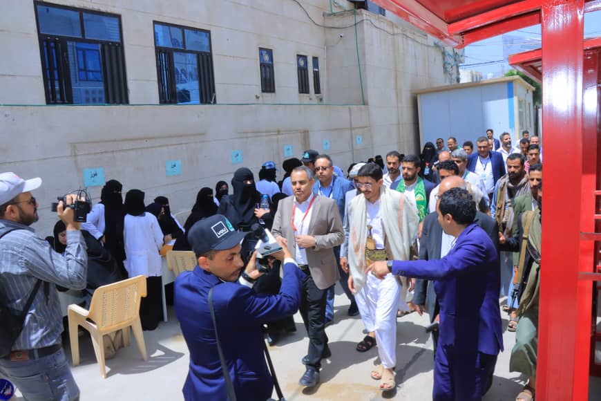 Outdoor event with a press crew filming as a man in a suit interacts with attendees.