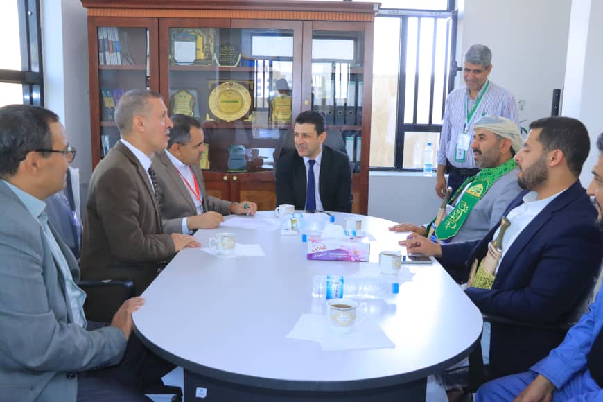 Eight men in suits and traditional attire sit around a white oval conference table, engaged in a discussion; a display cabinet with awards is in the background.