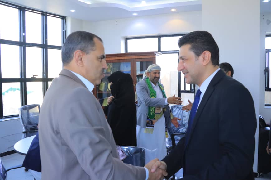 Two men in suits shake hands in a bright office while others greet in the background, suggesting a formal meeting or greeting.