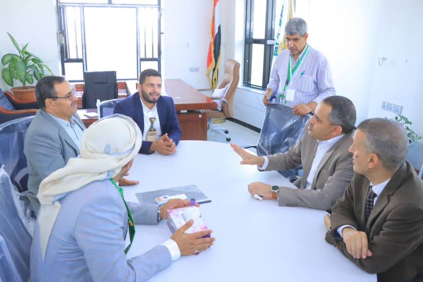 Six professionals in a meeting around a white round table in a bright office; one man stands with a lanyard as others listen and converse.