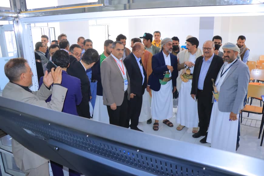 Group of men in suits and traditional robes on a guided tour inside a bright facility with orange chairs in the background.
