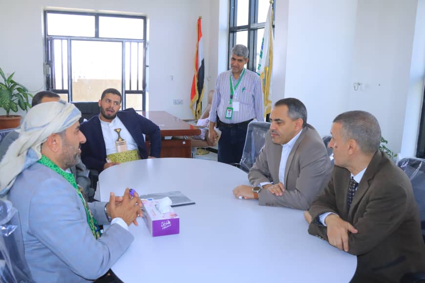 Group of men in suits seated around a round table in a conference room, one man standing with a badge near flags in the background.