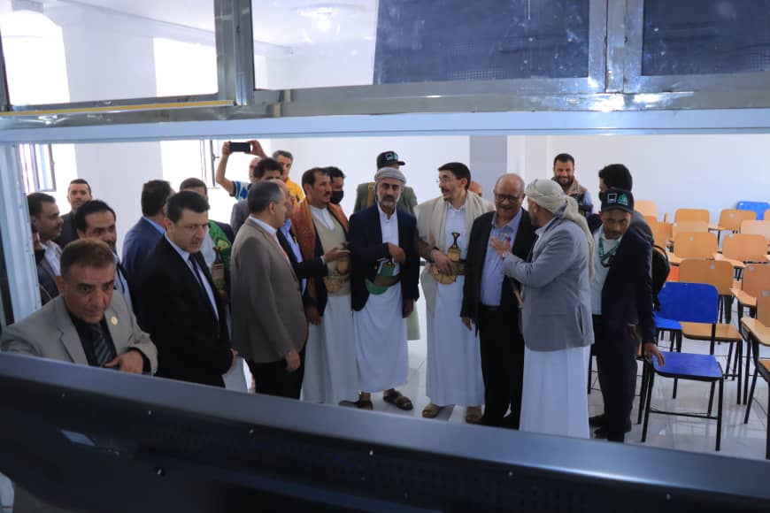 Group of men in traditional and business attire gathering around a large screen in a bright classroom, watching a demonstration.