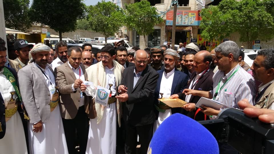 Group of men in traditional robes standing outdoors as a man in a suit hands something to another, with a blue microphone in the foreground.