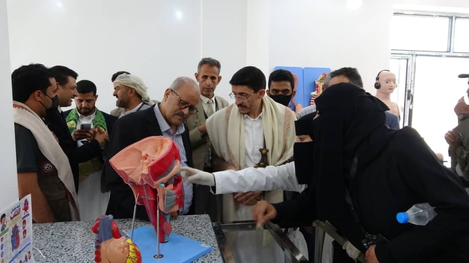 Group of people observing a medical demonstration: a woman in a black hijab points to a large anatomical heart model on a blue base placed on a counter