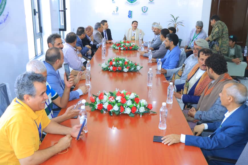 A formal meeting around a long wooden conference table with about twenty men on both sides, flower centerpieces, and water bottles.