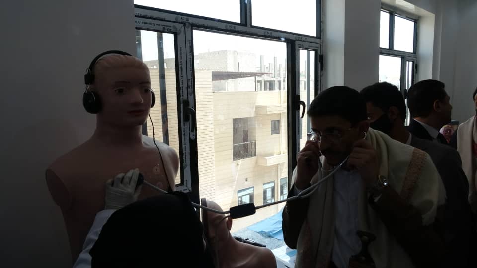 Medical training scene with a life-size mannequin wearing headphones while a trainee uses a stethoscope on its chest near a window.
