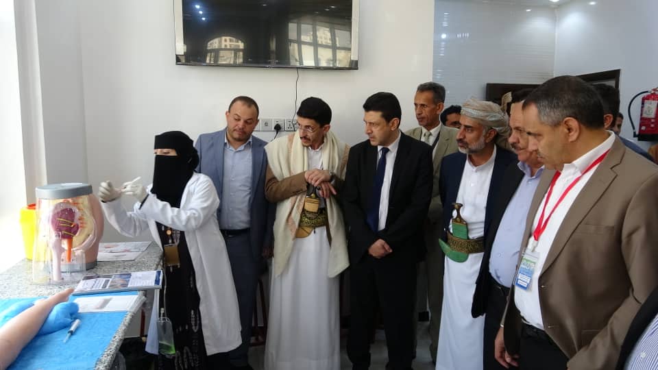 A diverse group of men in business and traditional attire observing a medical demonstration by a woman in a white coat and hijab at a lab counter with a transparent organ model on display.
