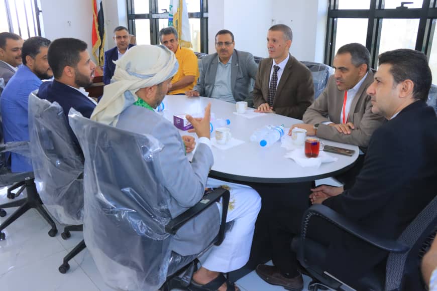 Nine people around a round table in an office, with flags visible in the background, engaged in a discussion over drinks and papers.
