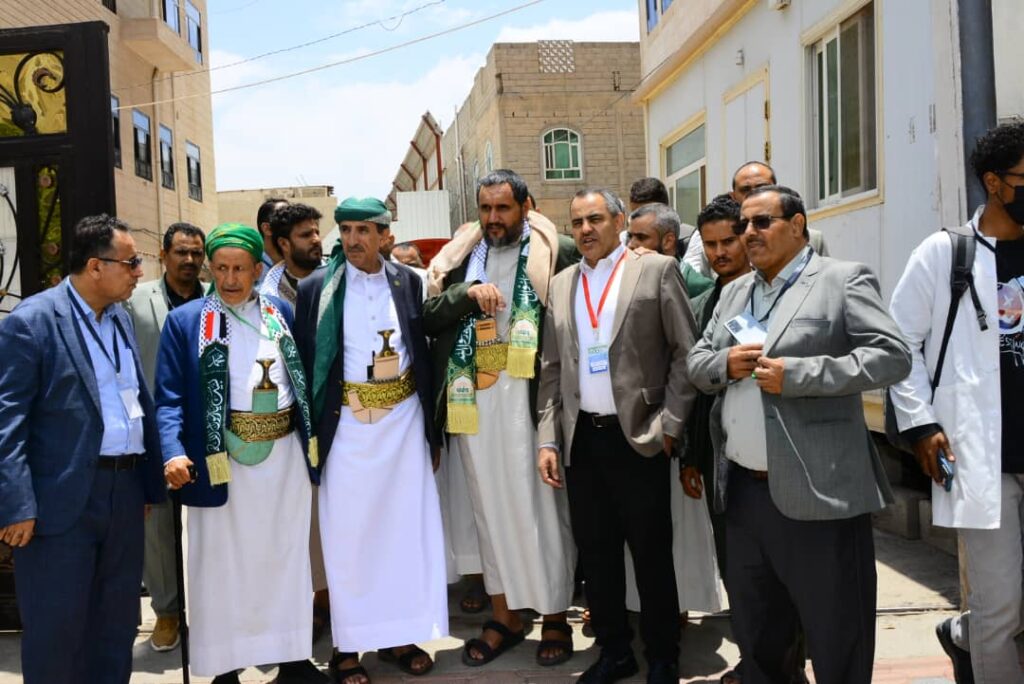 Group of men in traditional Yemeni attire with green scarves and ceremonial belts standing together on a sunny street.