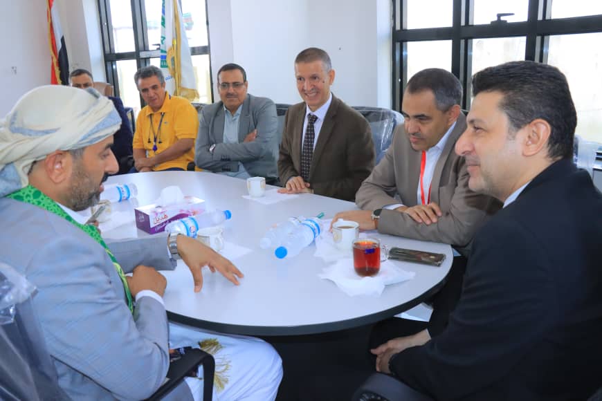 Group of men in a business meeting around a round table, discussing topics with drinks and papers on the table in a bright office setting.