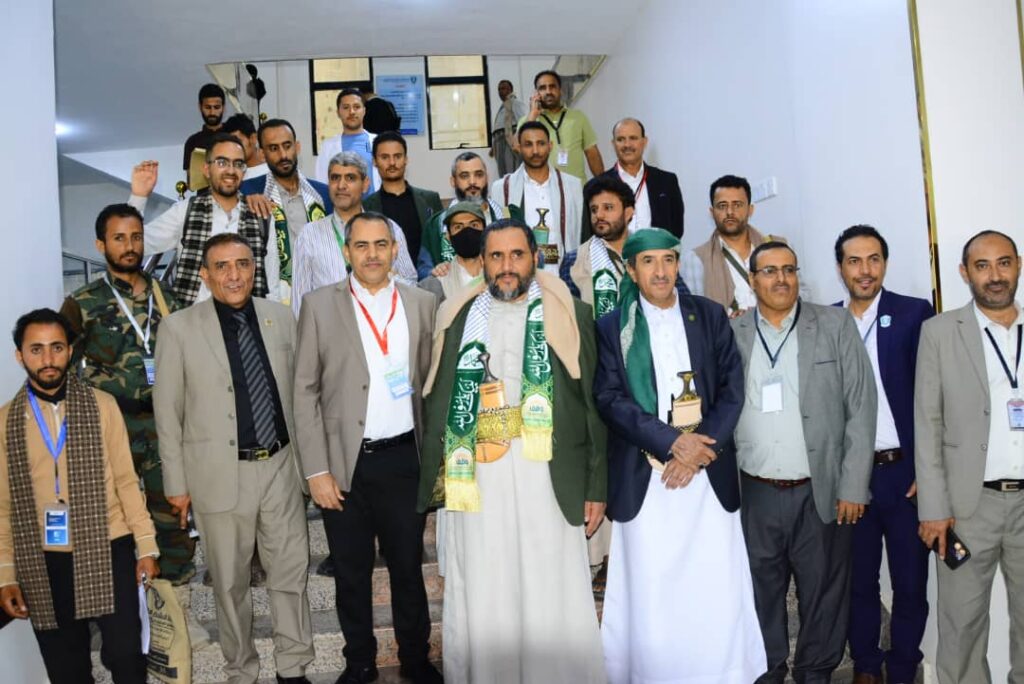 A large group of men posing on a stairwell for a formal group photo at an event, some wearing lanyards, traditional robes and green scarves.
