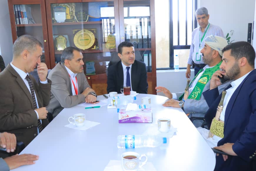 Group of men in suits seated around a white conference table in discussion, one man in a green ceremonial vest talking to colleagues nearby in an office setting.
