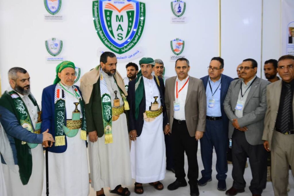 Group of men in traditional robes and scarves posing at a formal event with a large MAS logo in the background.