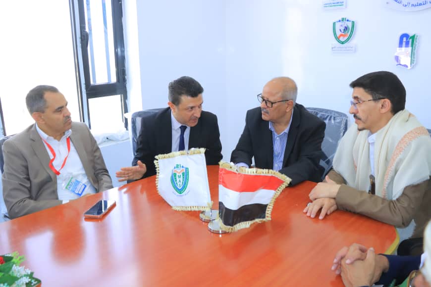 Four men in suits sit around a wooden conference table, discussing, with two decorative flags on the table (one featuring a shield emblem).