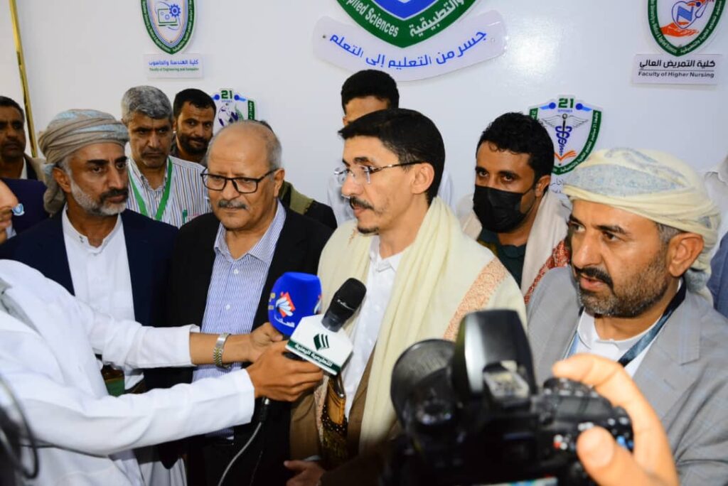 Group of men at a press conference; reporter holds two microphones toward a central speaker wearing a cream scarf, with cameras recording nearby and Arabic banners in the background.