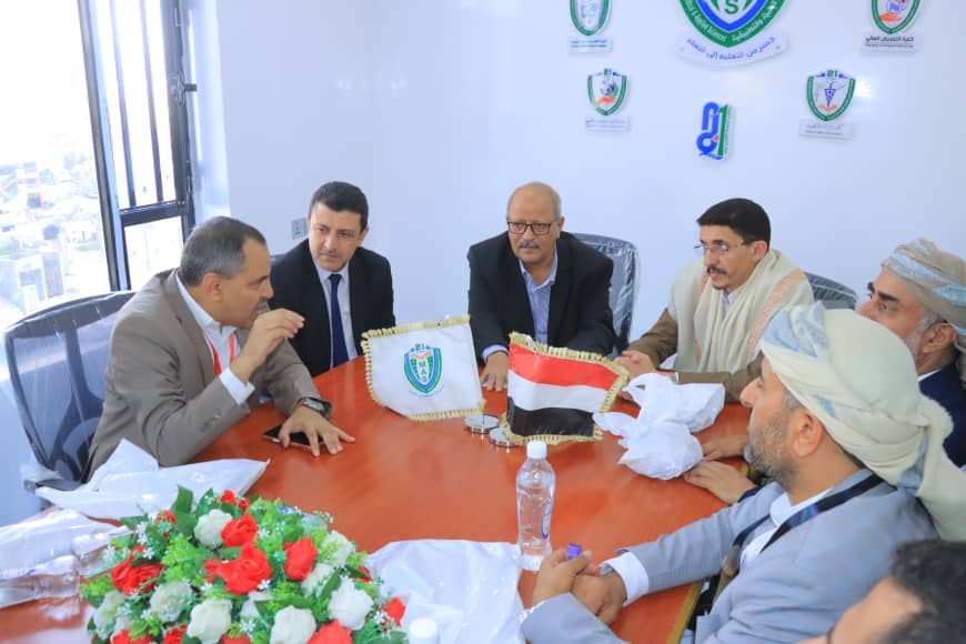 Group of men in suits and traditional dress sit around a conference table in a bright office, engaged in a formal discussion; small flags on the table suggest a diplomatic or organizational meeting.